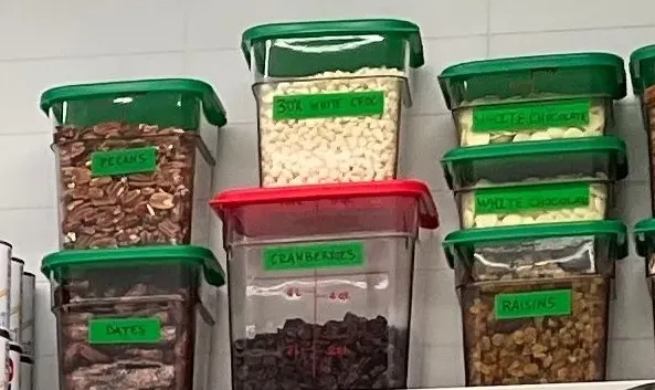 Square storage containers with green and red lids on the shelf, labelled pecans, dates, cranberries, white chocolate chips and raisins